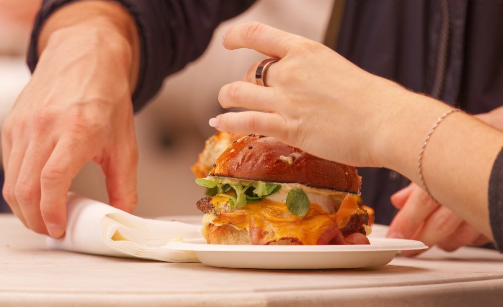 Close-up of two people sharing a gourmet burger with melted cheese, fresh greens, and a brioche bun at an outdoor food market.
