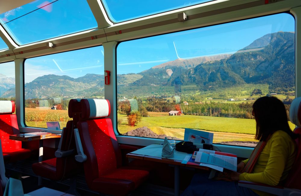 A woman relaxes in a panoramic train carriage, gazing out at sun-drenched alpine fields and mountains through large windows, as sunlight pours into the cozy red-seated cabin.