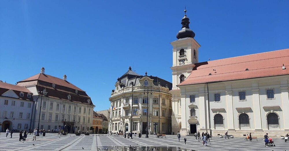 A sunlit square featuring historic buildings with varying architectural styles, bustling with people under a clear blue sky.
