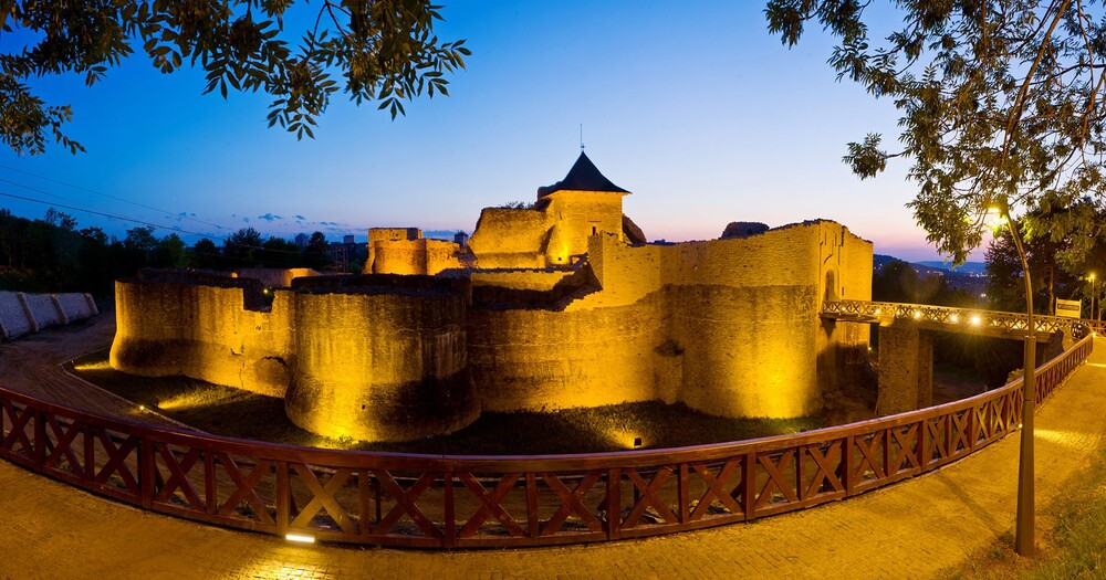 Illuminated medieval castle at twilight, surrounded by trees, with a wooden walkway and a clear sky transitioning to night.