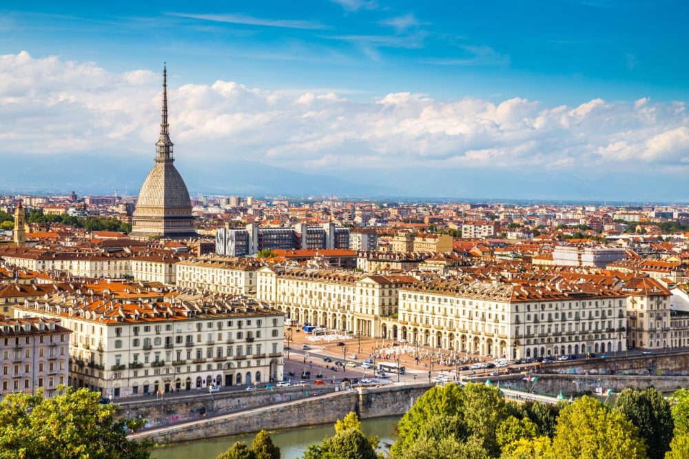 A panoramic view of Turin featuring the iconic Mole Antonelliana, overlooking historic buildings and the Po River beneath a blue sky.