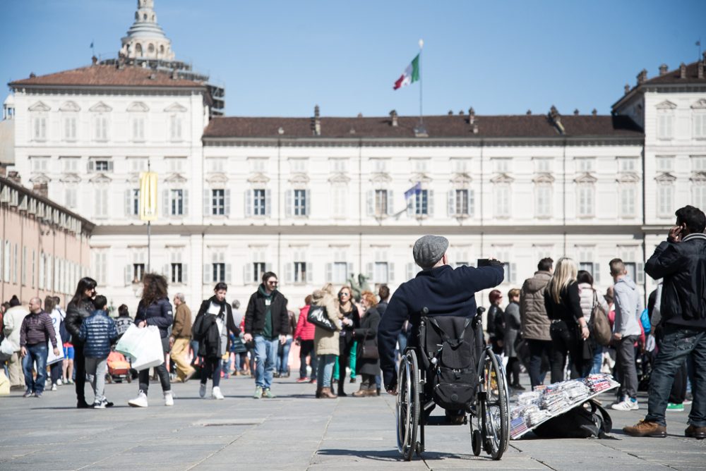 A man in a wheelchair takes a selfie amidst a busy plaza filled with people, with historic architecture in the background.
