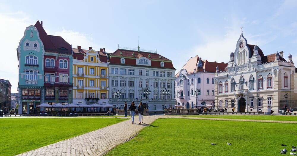 Colorful historic buildings line a grassy square, with people walking on a path under a bright blue sky.