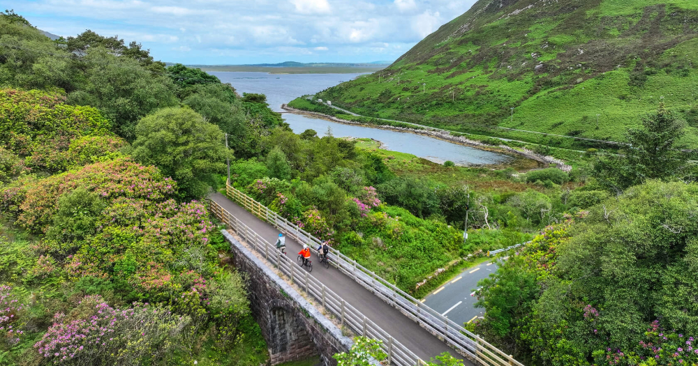 Two cyclists ride on a winding road beside vibrant greenery and a serene river, with a hilly landscape under a cloudy sky.
