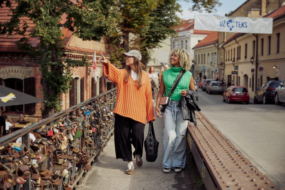 Two women walking across the love lock bridge leading to Užupis in Vilnius, Lithuania, on a sunny day.
