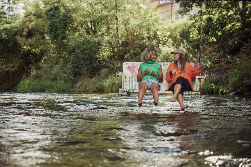 Two friends sitting barefoot on a swing over the Vilnia River in Vilnius, laughing and enjoying a summer day surrounded by greenery.