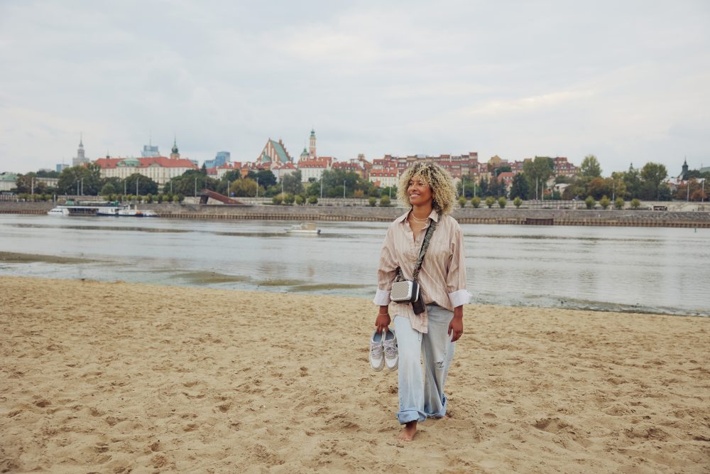Woman walking barefoot on a sandy beach by the Vistula River with Warsaw’s Old Town skyline in the distance.