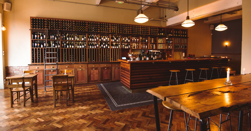A cozy wine bar interior featuring a wooden counter, shelves filled with bottles, and rustic tables under warm lighting.