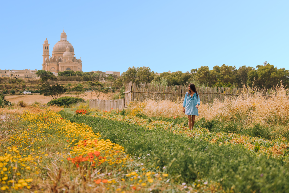 A woman in a blue dress strolls through a vibrant flower field with a historic dome building in the background under a clear sky.