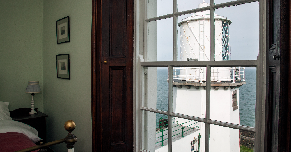 A view of a white lighthouse seen through a window from a cosy room.